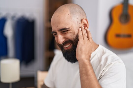 Young bald man suffering for ear pain sitting on bed at bedroom