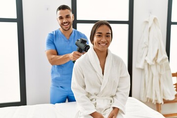 Latin man and woman wearing physiotherapy uniform massaging neck using percussion gun at beauty center