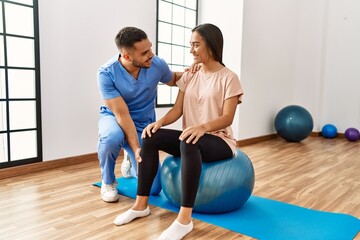 Latin man and woman wearing physiotherapist uniform having rehab session using fit ball at rehab center