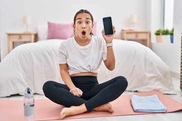 Young south asian woman doing yoga mat holding smartphone showing screen scared and amazed with...