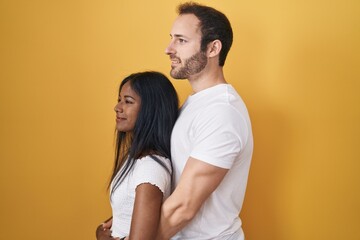 Interracial couple standing over yellow background looking to side, relax profile pose with natural face and confident smile.