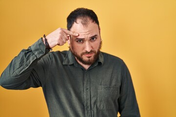 Plus size hispanic man with beard standing over yellow background pointing unhappy to pimple on forehead, ugly infection of blackhead. acne and skin problem