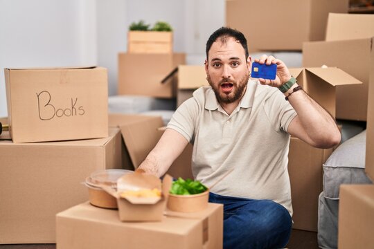 Plus size hispanic man with beard moving to a new home holding credit card scared and amazed with open mouth for surprise, disbelief face