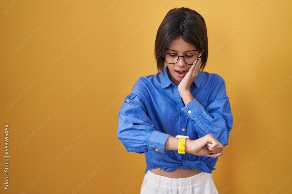 Wall mural young girl standing over yellow background looking at the watch time worried, afraid of getting late