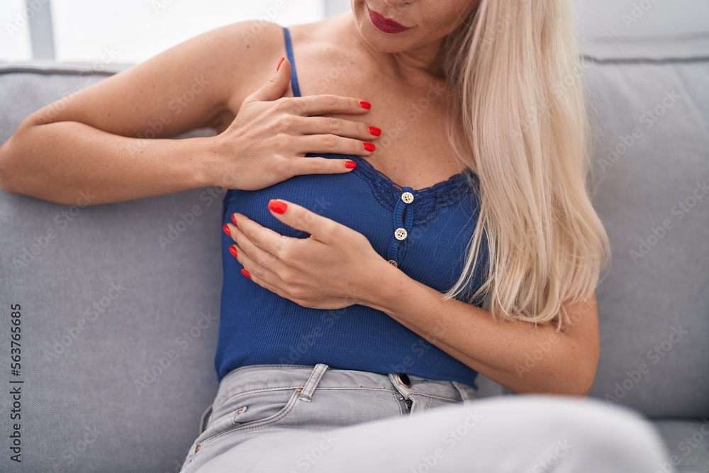 Sticker Young blonde woman sitting on sofa examining breast with hands at home