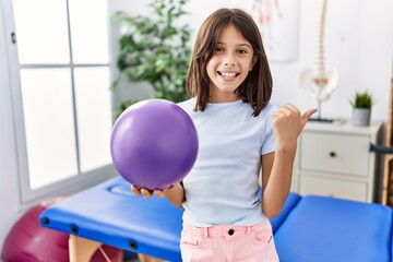 Young hispanic girl holding small rehabilitation ball pointing thumb up to the side smiling happy...