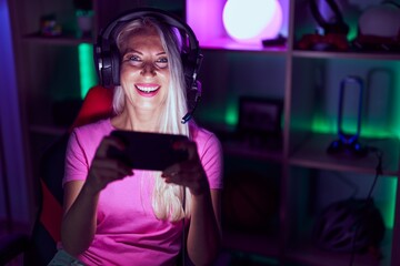 Obraz na płótnie Canvas Young blonde woman streamer playing video game by smartphone at gaming room