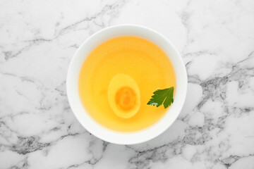 Delicious bouillon with egg and parsley in bowl on white marble table, top view
