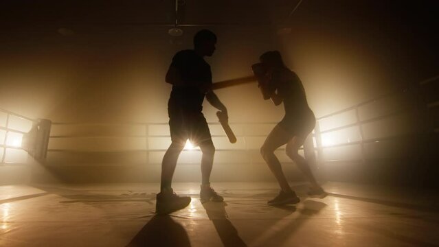 Close-up view of female athlete training in boxing club. Silhouettes of man and woman having intensive workout indoors. High quality 4k footage in golden orange yellow foggy light