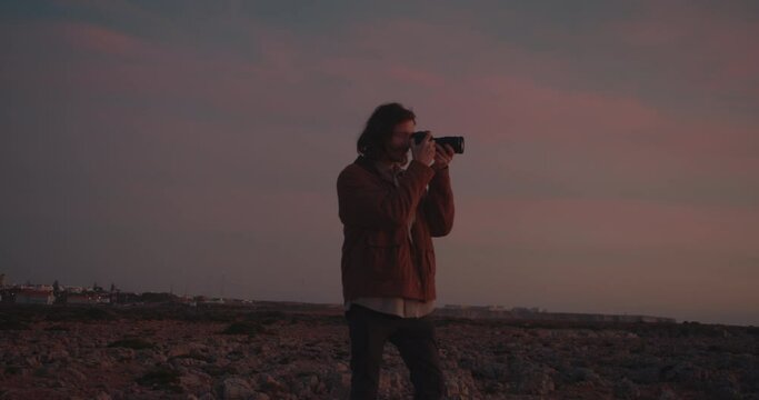 Long haired male photographer taking pictures and enjoying the scenery during sunset and blue hour on the coastline, cliffs of the Algarve, Portugal