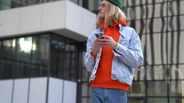 Stylish hipster guy holding smartphone connecting to public internet wifi while waiting friends outdoors. Young millennial man standing alone on city street using taxi app cellphone. Urban living