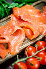 Slices of salted salmon with greens and tomatoes on a cutting board. 