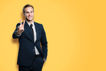 Excited businessman in black confident suit showing two fingers or victory sign gesture, on vivid yellow background. Handsome happy man. Copy space for ad, slogan or text.