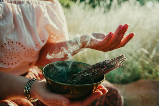 Woman sitting in a field of straw with an ornate bowl with a smudge stick burning and the smoke blowing over her hand.