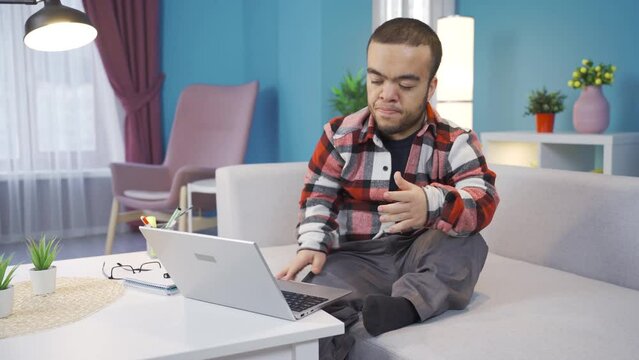 Frustrated exhausted dwarf young man working from home at computer.
Young disabled dwarf man typing on laptop and reading bad news online and getting angry and upset.
