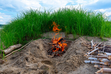 A campfire burns on a bright day in the sand next to tall green grass under blue clouds and birch firewood nearby.