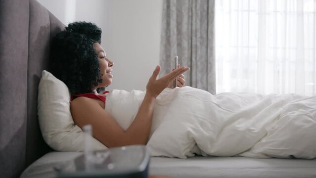 Side view bedroom apartment, beautiful African American woman using smartphone in bed at morning. Smiling girl on video call with friends of family. Slow motion wireless connection and communication