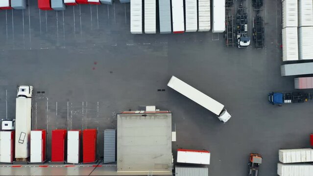 Semi-truck is driving along a warehouse with a lot of cargo containers standing at ramps for unloading and loading goods in the logistics park. Aerial top down view