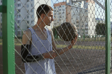 Serious male basketball player standing near net fence on urban playground in city 