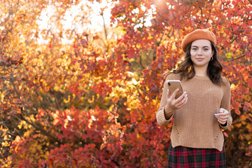 Hello october. Happy stylish woman in orange beret using smartphone applications among autumnal foliage outside on the city park in autumn.