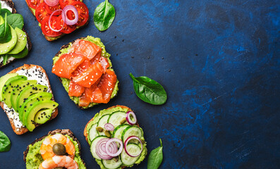 Avocado sandwiches or toast set: with salmon, shrimps, tomatoes, cucumbers, soft cheese and spinach, crushed cashew nuts and sesame seeds, blue table background, top view