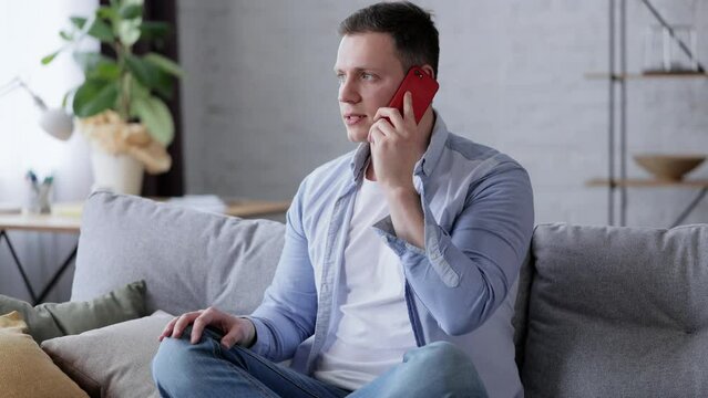 Nervous man speaking on smartphone. Young Caucasian Guy sits on sofa in house in cozy living room speaking on cellphone in bad mood. Portrait of male at home speaking emotionally on smartphone.