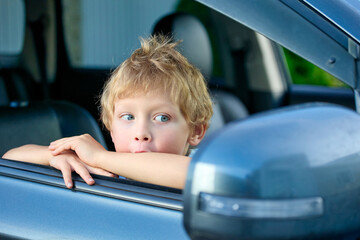 Small child looks in surprise at the side mirror of the car sitting in the passenger seat and leaning out of the window. The boy watches what is happening in the car mirror