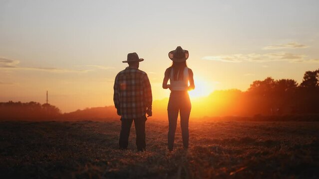Back view of two farmers: man and woman walking along plowed field on red sunset. Friends relax in evening walk on outdoor nature. People talking and gesture. Rural scene. Agronomy and agricultural.