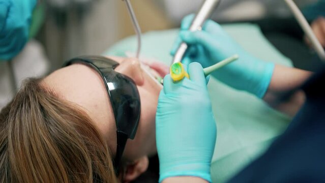 Woman lying on chair on cabinet tooth clinic with mouth dilator and protective goggles. Dentist in glasses with magnifying glass holding medical equipment. UV lamp for whitening or caries treatment.