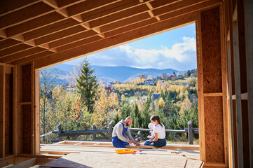 Father, mother and son building wooden frame house in the Scandinavian style barnhouse. Toddler boy helping his daddy, while woman looking for them on construction site. View from inside a house.