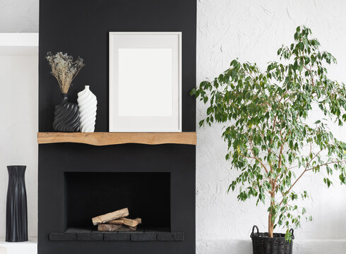 Blank picture frame mockup on black wall. White living room design. View of modern scandinavian style interior with fireplace, trendy vase and plant. Home staging and minimalism concept