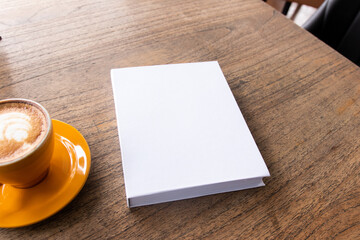 A white colored hardcover book with a wooden textured table as the background and a coffee near it