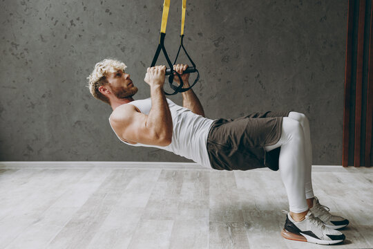 Full body sideways young strong sporty athletic sportsman man wearing white tank shirt black shorts doing exercises with trx loops looking camera warm up training indoor at gym. Workout sport concept.