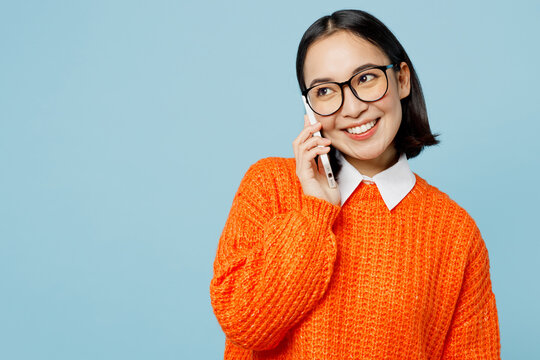 Young woman of Asian ethnicity in orange sweater glasses talk speak on mobile cell phone conducting pleasant conversation isolated on plain pastel light blue cyan background People lifestyle concept