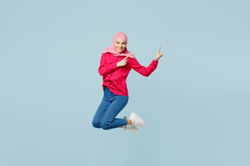 Full body side view young arabian asian muslim woman in pink abaya hijab jump high point finger finger aside area isolated on plain light blue cyan background studio portrait People uae islam concept.