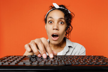 Close up young surprised woman of African American ethnicity wears grey shirt headband typing on pc computer keyboard look camera with opened mouth isolated on plain orange background studio portrait.