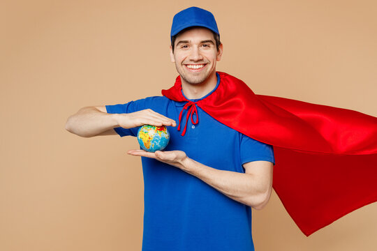 Delivery guy employee man wear blue cap t-shirt uniform workwear super hero red coat work dealer courier hold cardboard box Earth world globe isolated on plain light beige background. Service concept.