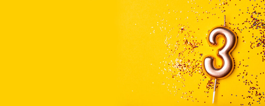 Gold candle in the form of number three on yellow background with confetti.