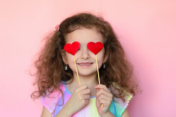 Portrait little cute girl holding two red hearts in hands near eyes on pink background. creative...