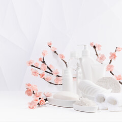 Beauty products for body, skin care in white bottles, branch of pink sakura flowers, bath toiletry on table in modern soft light white interior in minimalist geometric japan style, copy space, square.
