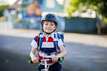 Fototapeta na wymiar Active school kid boy in safety helmet riding with his scooter in the city with backpack on sunny day. Happy child biking on way to school. Safe way for kids outdoors to school.