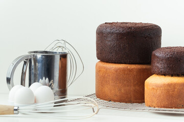 Freshly baked sponge biscuit on a pastry grid next to whisk, sieve for sifting flour and eggs. Brown chocolate and white vanilla sponge cake with professional confectioner utensils. White background