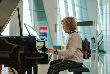 An adult woman with a backpack plays the piano at an airport while waiting for her flight to depart.