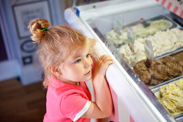 Cute little toddler girl choosing and buying ice cream in a cafe. Happy baby child looking at...