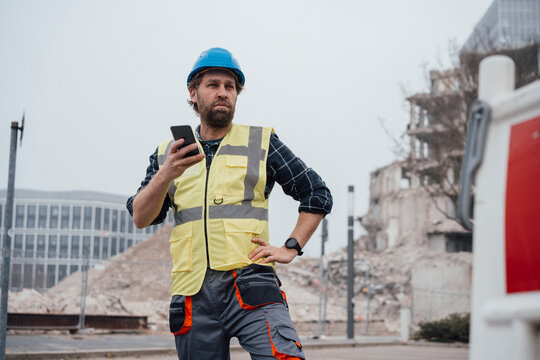 Worker Holding Smart Phone With Hand On Hip In Front Of Construction Site