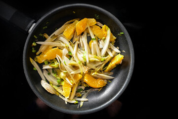 Slices of chicory and orange with spring onions in a black frying pan on the cooker to, vegetarian and vegan meal for a healthy low carb diet, high angle view from above, copy space