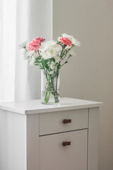 Fresh chrysanthemums and carnations in a glass vase on a white chest of drawers in a cozy bright room