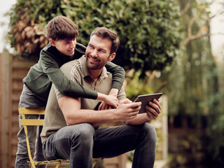 Son embraing father sitting in garden using digital tablet