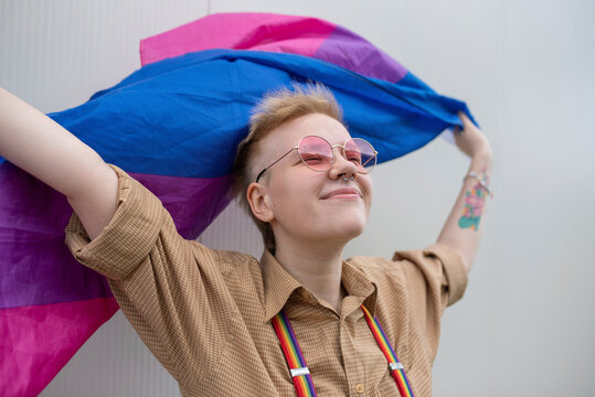 Smiling non-binary person holding pride flag in front of wall