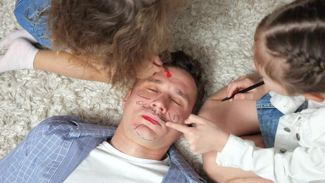 Girls siblings paint sleepy father face with bright markers. Obedient man sleeps and has nice dreams lying on floor with carpet upper view